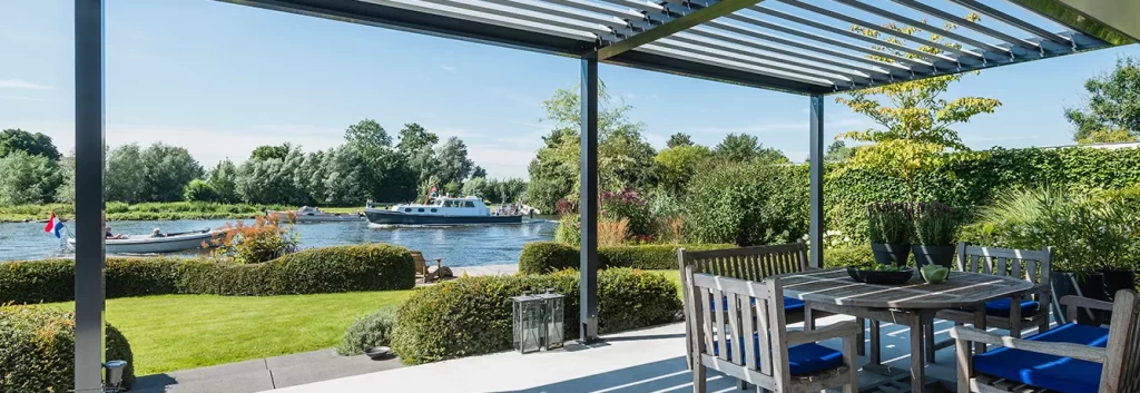 Riverside patio with shaded pergola, wooden table and chairs.